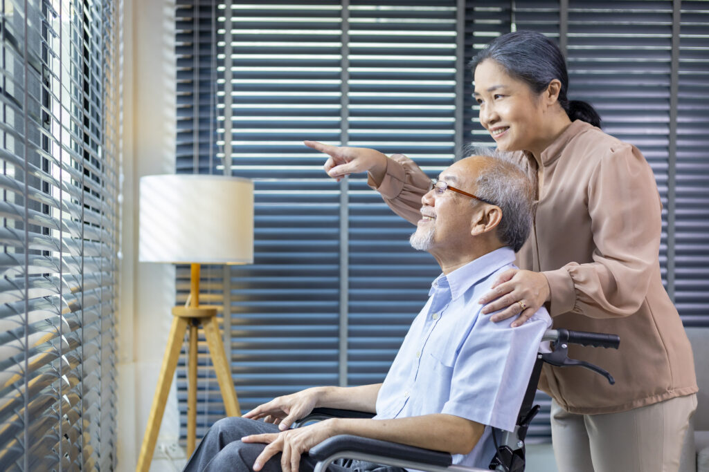 An older couple together beside a window in their home.